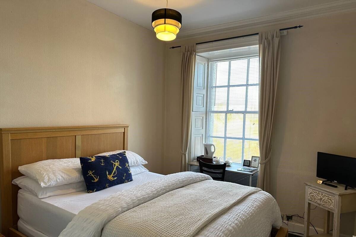 A neatly made bed with a wooden headboard, a bedside table with a kettle and glasses, and a television on a small stand.