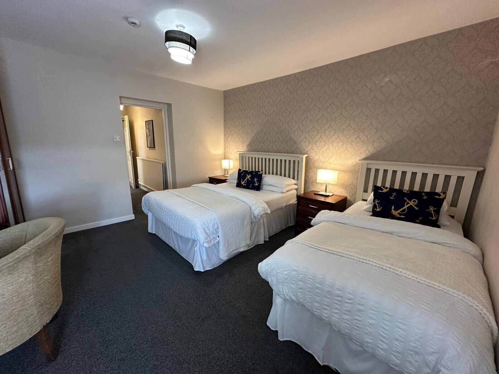 Hotellihuone, jossa on kaksi erillistä sänkyä, pieni yöpöytä lampun kanssa, tuoli ja ovi, joka johtaa toiseen huoneeseen.