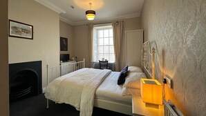 A bedroom with a bed, a bedside table with a lamp, a chair, a desk with a computer, a fireplace, and a window with curtains.