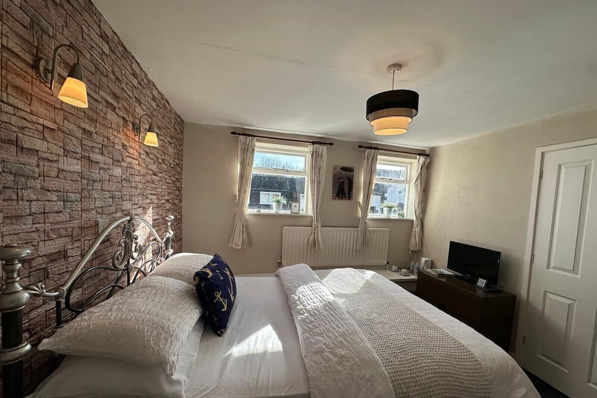 A bedroom with a brick wall, a bed with white bedding, a nightstand, a television, and a window with curtains.