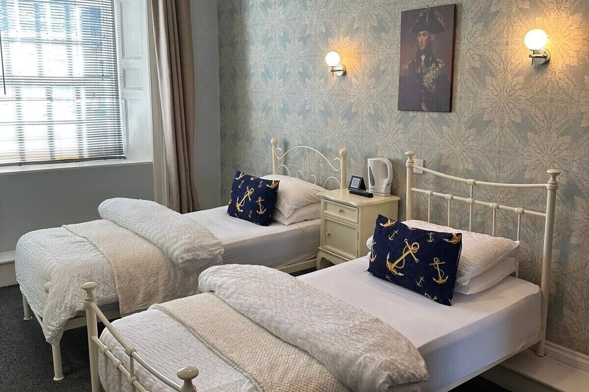 A bedroom with two beds, a window with blinds, a nightstand, a lamp, and a wall-mounted picture.