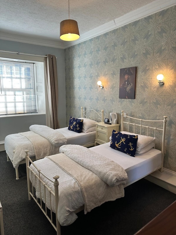 A bedroom with two beds, a window with blinds, a nightstand, a lamp, and a wall-mounted picture.