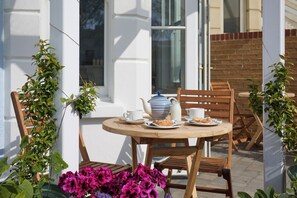 A round wooden table with chairs, a white teapot, a milk jug, and plates with pastries, set against a white wall with a window and a brick wall in the background.