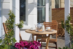 A round wooden table with chairs, a white teapot, a milk jug, and plates with pastries, set against a white wall with a window and a brick wall in the background.