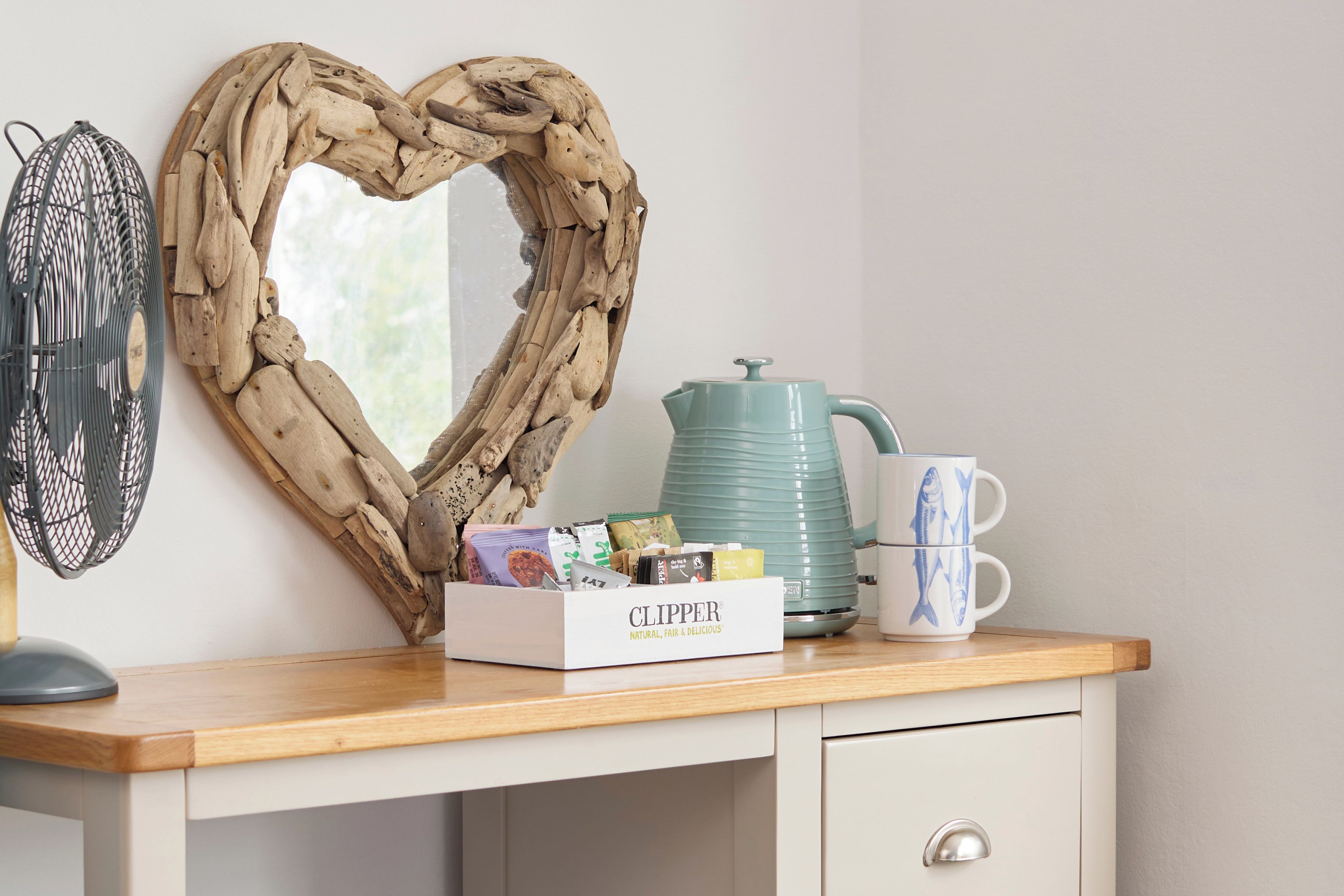 Un escritorio de madera con un espejo con forma de corazón, una tetera, una caja de té Clipper y una taza con motivos de peces.