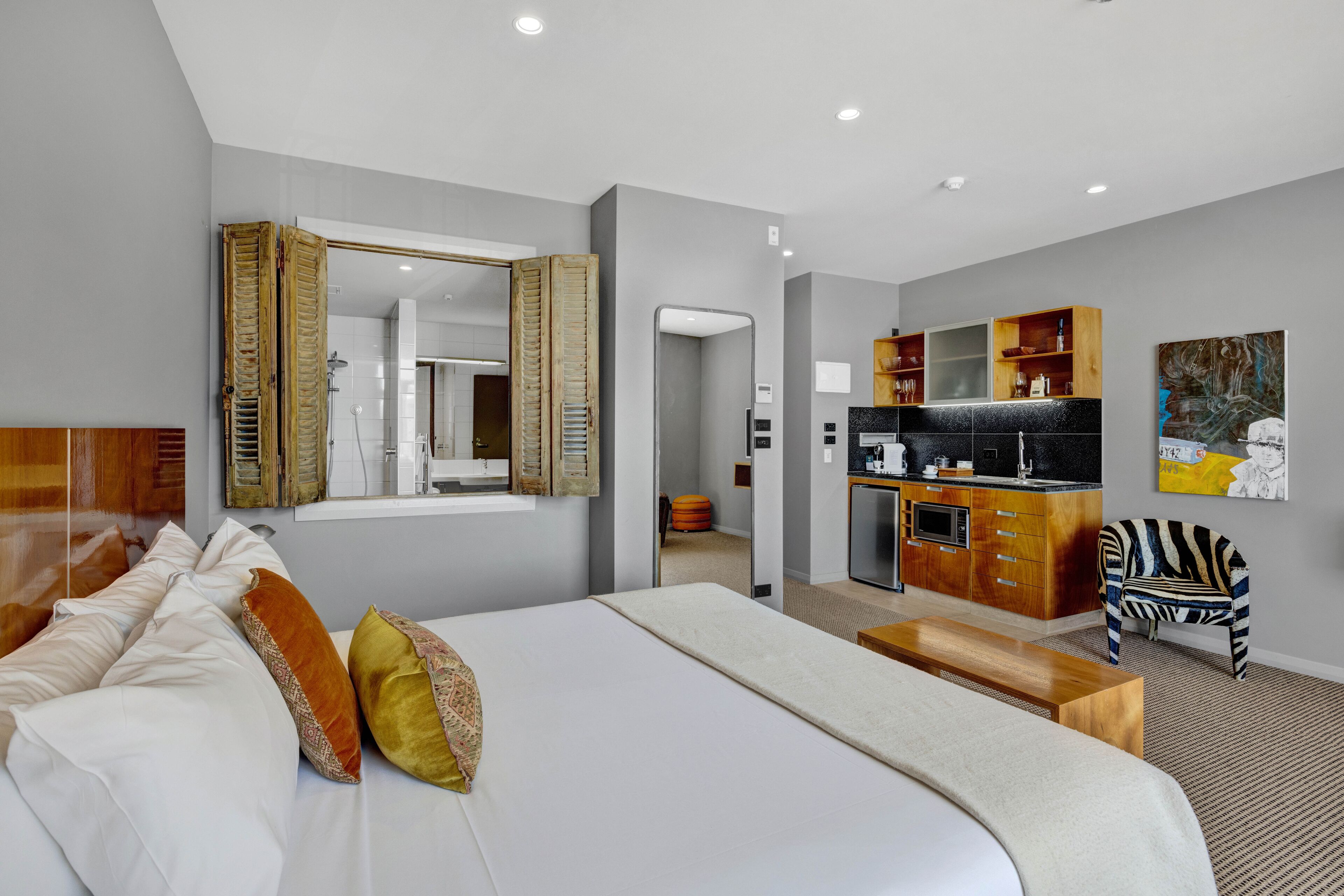 A modern hotel room with a large bed, a wooden bench, a kitchenette, and a zebra-striped chair.