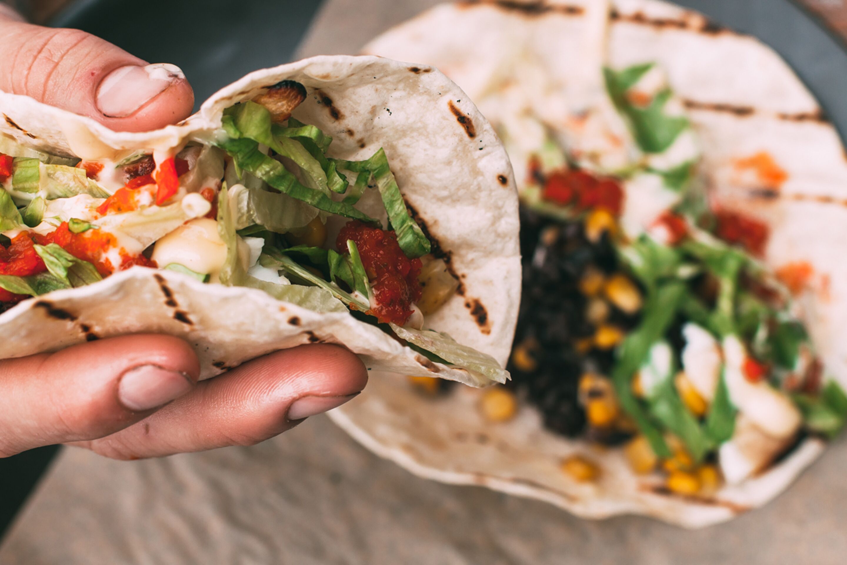 Uma pessoa segurando um burrito com recheios à vista, como alface, tomates e milho.
