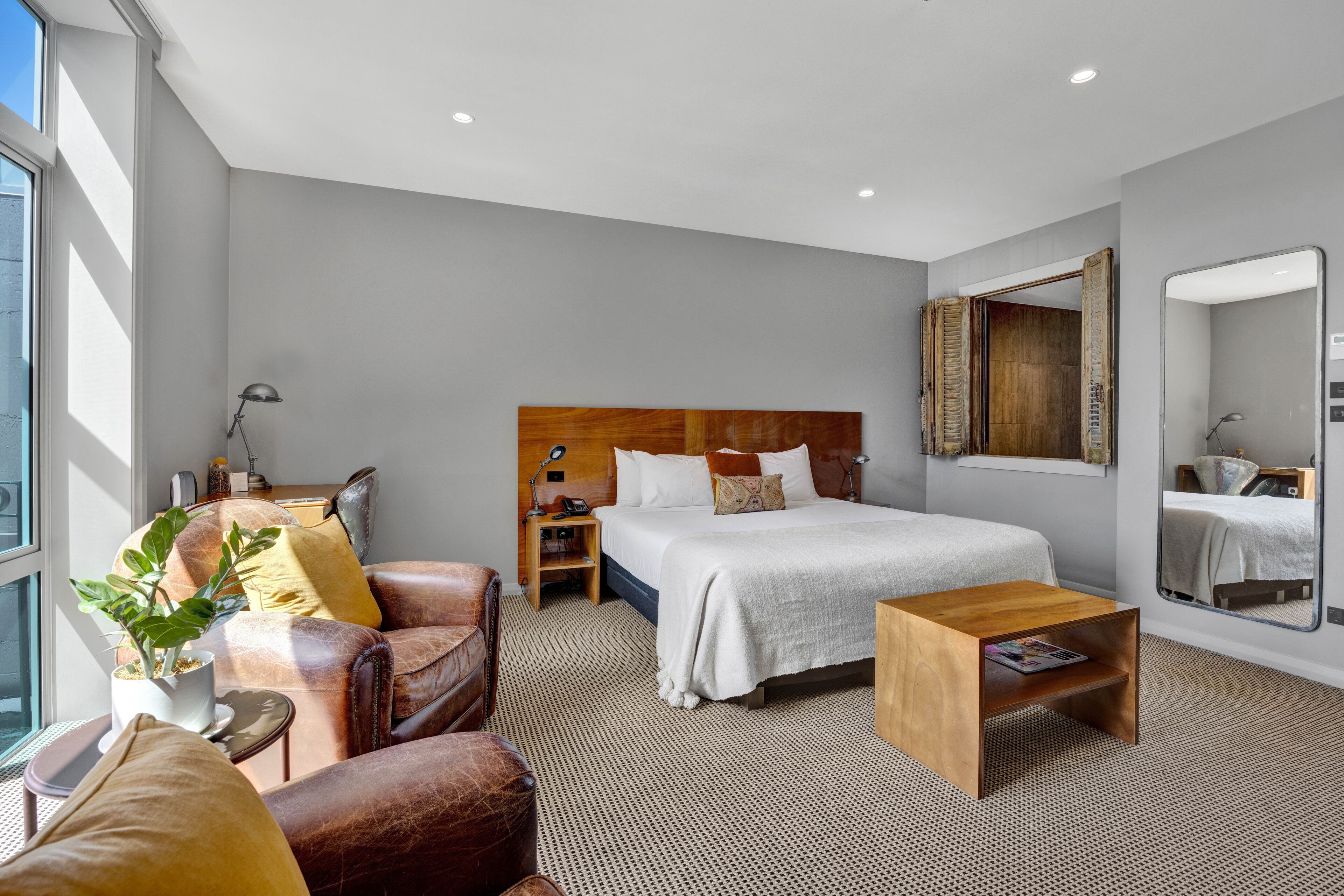 Ein modernes Hotelzimmer mit einem großen Bett, einem Holzkopfende, einem kleinen Nachttisch, einem Spiegel und einer Zimmerpflanze.