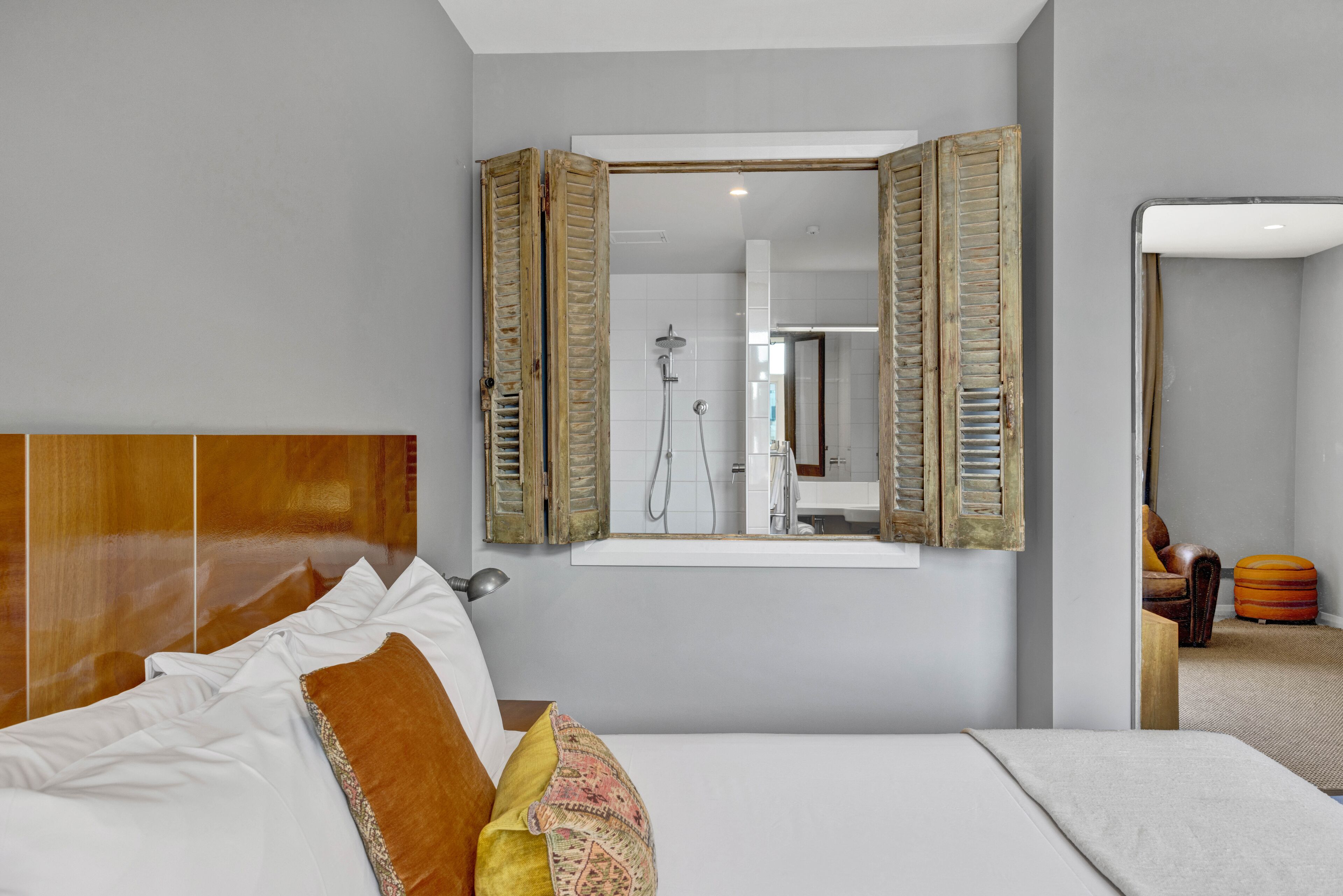 A hotel room with a large bed, a wooden headboard, a mirror with shutters, a shower area, and a small seating area with a chair and ottoman.
