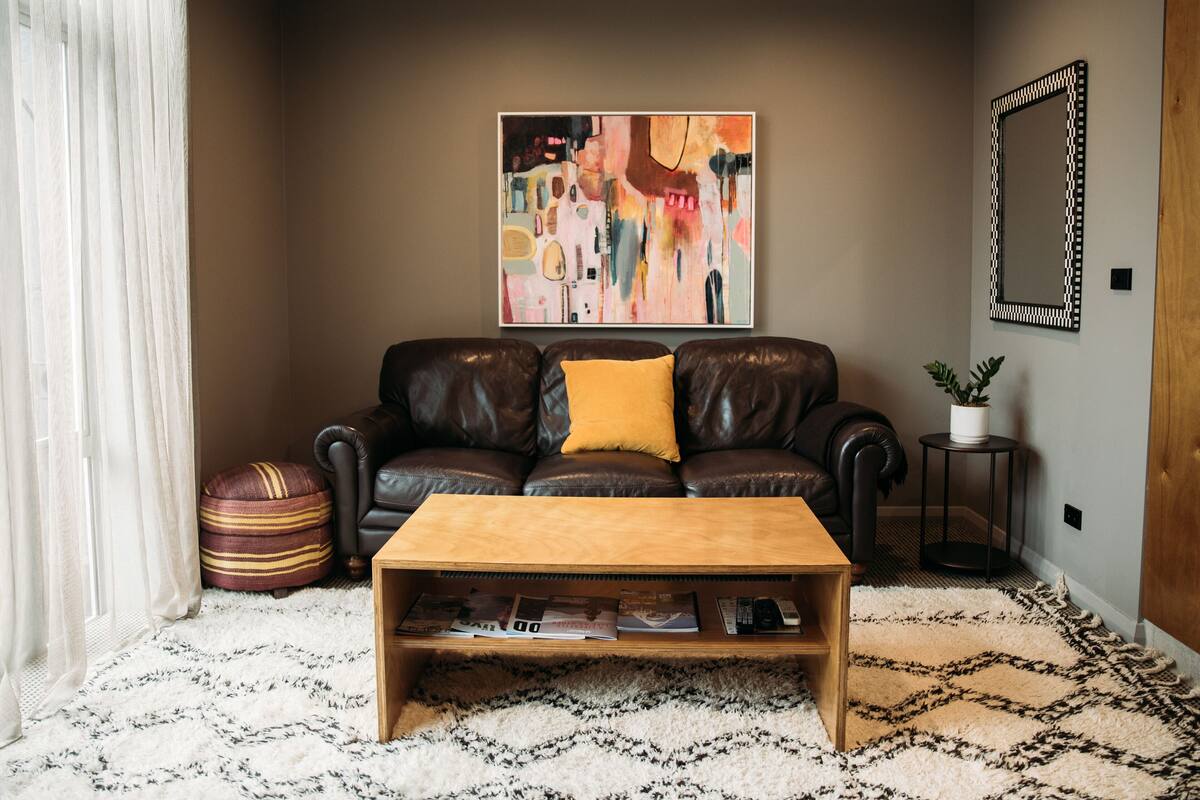 Un salon avec un canapé en cuir marron, une table basse en bois, un tableau abstrait aux couleurs vives, un miroir et un tapis à motifs.