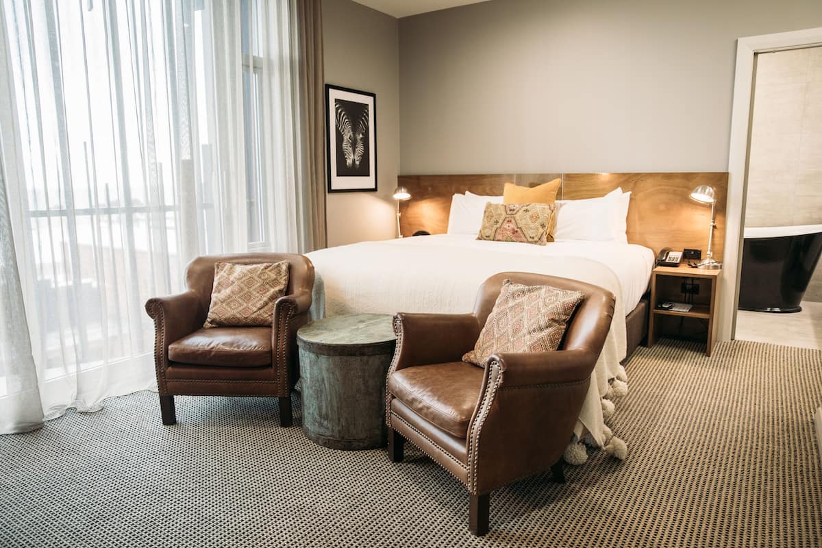 Une chambre d’hôtel avec un grand lit, deux fauteuils en cuir, une petite table ronde et une table de chevet avec une lampe.