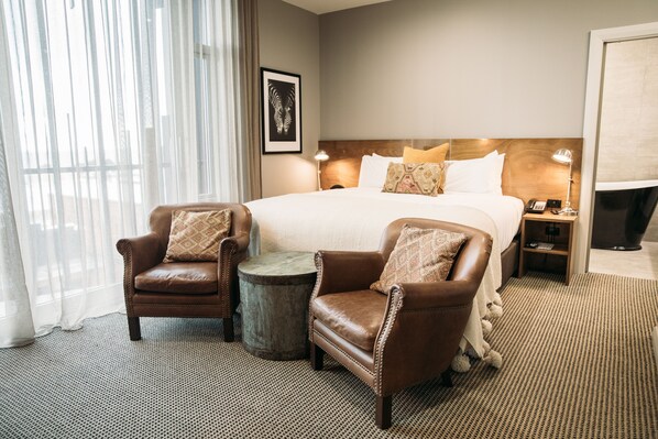A hotel room with a large bed, two leather armchairs, a small round table, and a nightstand with a lamp.