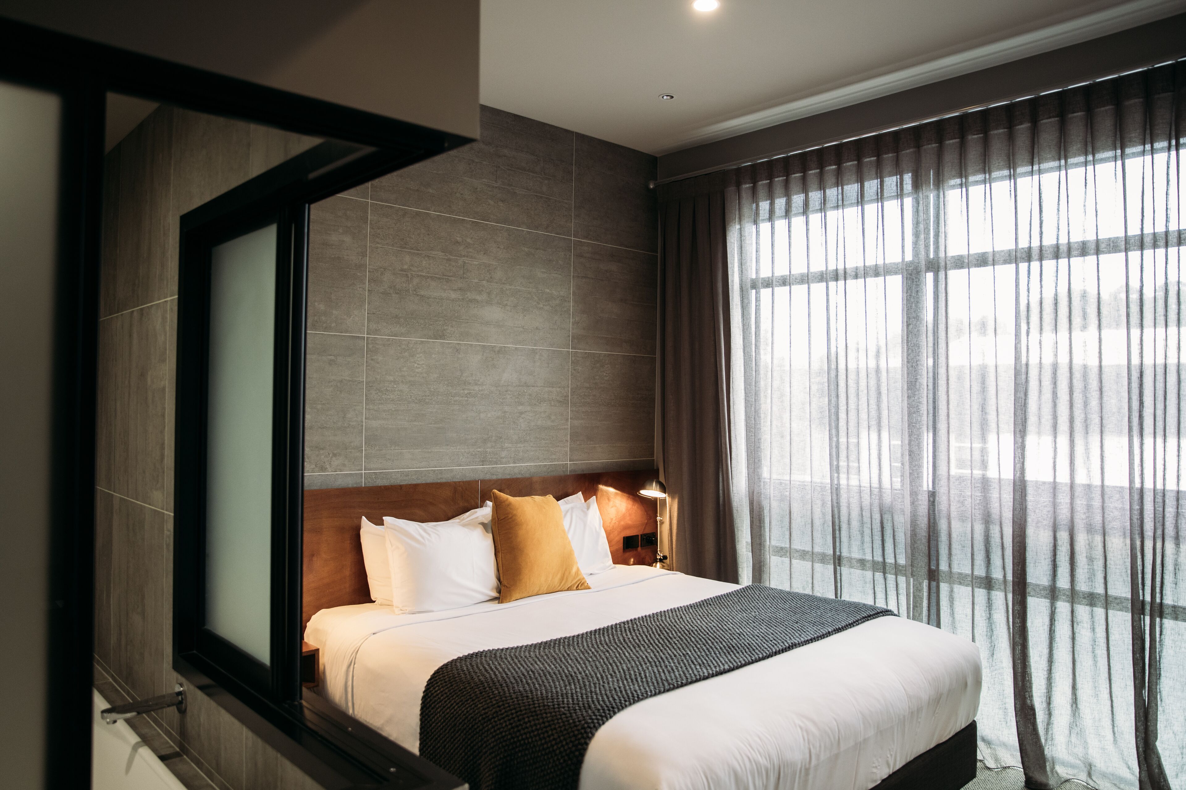Une chambre d’hôtel moderne avec un grand lit, une table de chevet, une fenêtre avec des rideaux légers et une vue sur la ville.