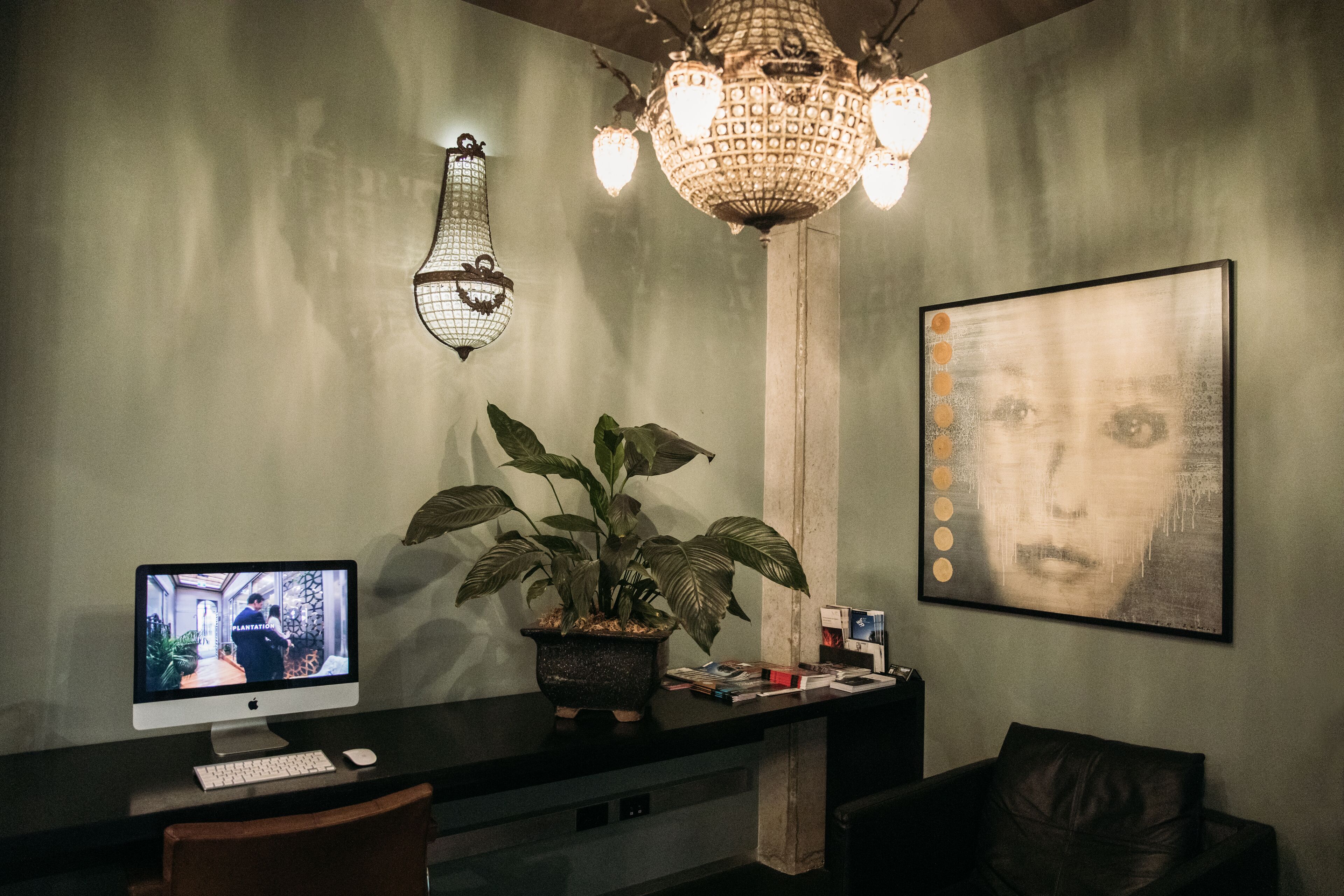 A reception desk with two monitors, a potted plant, a chandelier, and a framed artwork on the wall.