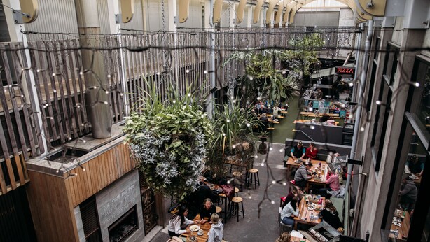 Un comedor interior con mesas y sillas de madera, un puesto de pizzas y plantas colgantes.