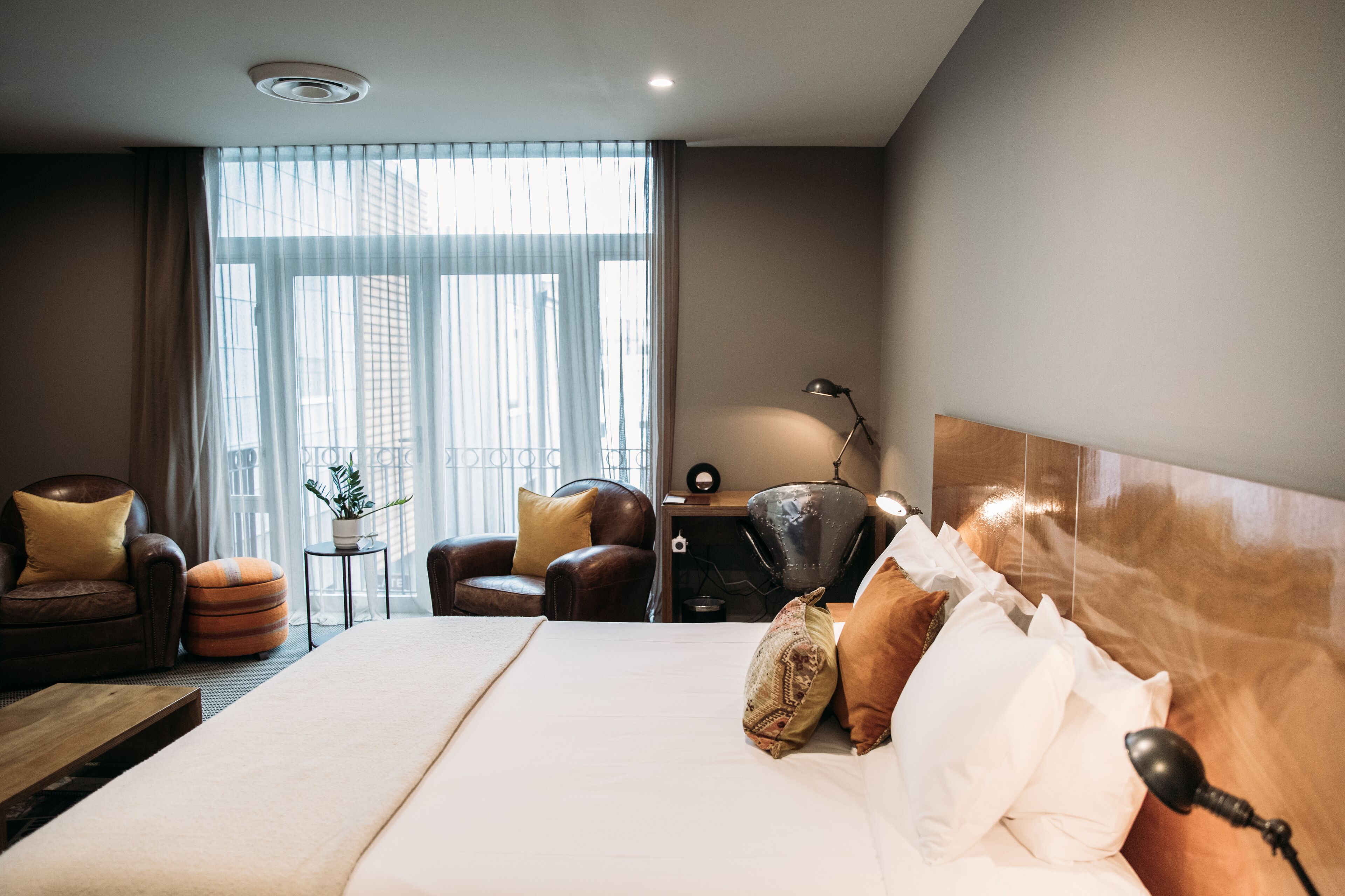 A hotel room with a large bed, two armchairs, a small table with a lamp, and a view of the city through large windows.
