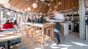 Un restaurante moderno con mesas de madera y taburetes de bar, zona de cocina y grandes ventanales que dejan entrar luz natural.