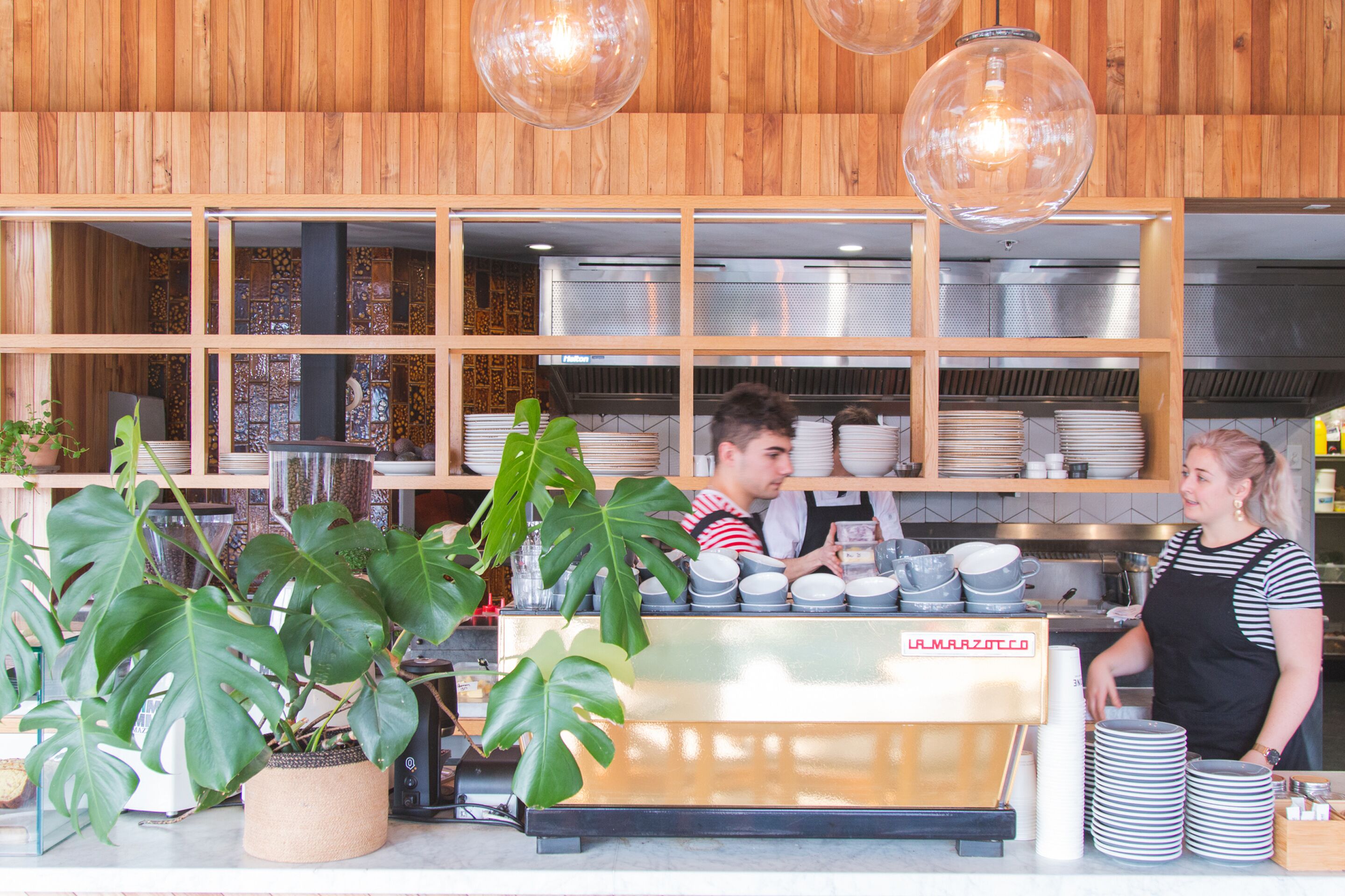 Interior de una cafetería con mostrador, máquina de café y estantes llenos de vajilla.