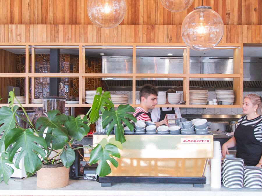 Interior de una cafetería con mostrador, máquina de café y estantes llenos de vajilla.