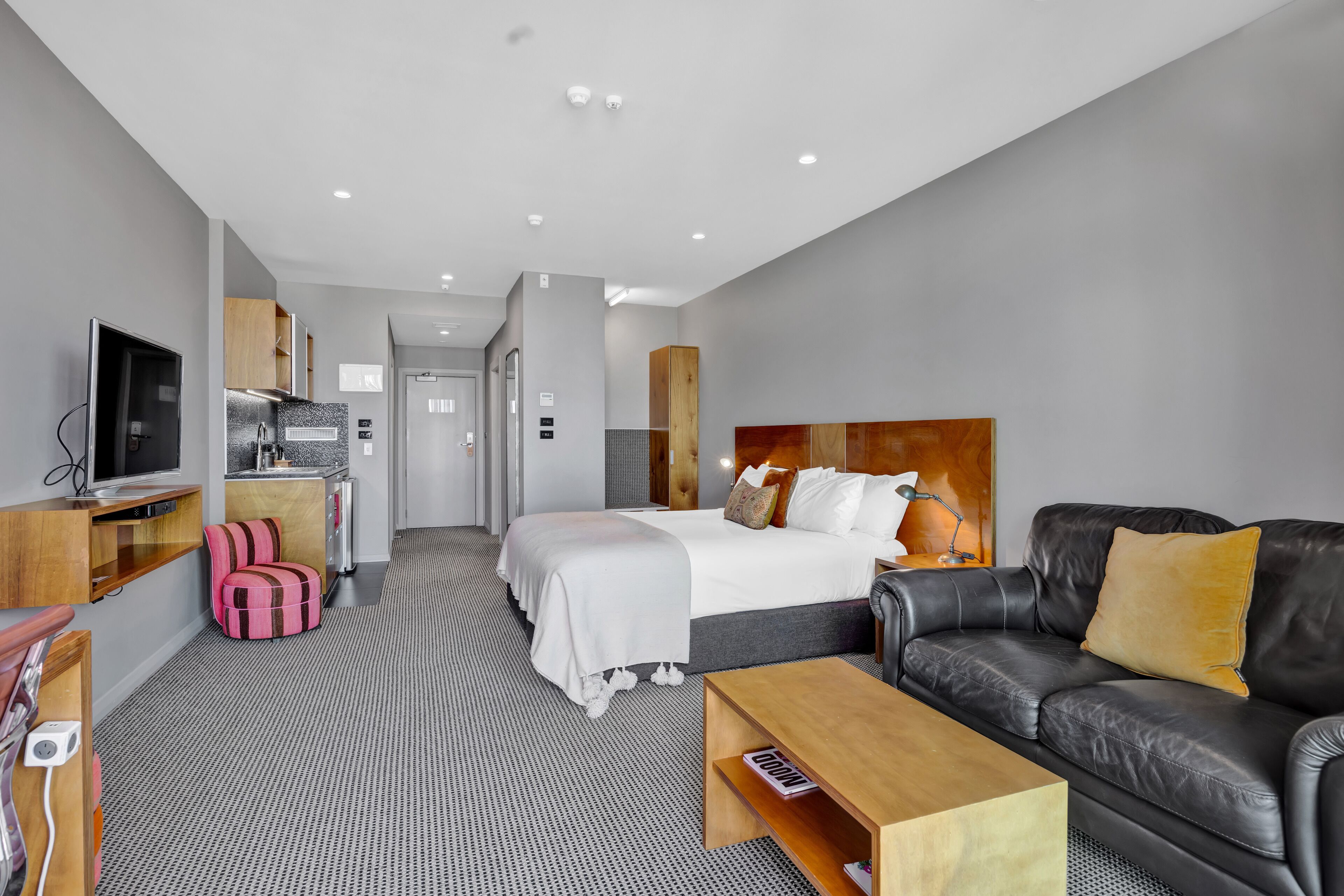 A modern hotel room with a bed, a sofa, a TV, a desk, and a kitchenette.