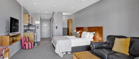 A modern hotel room with a bed, a sofa, a TV, a desk, and a kitchenette.