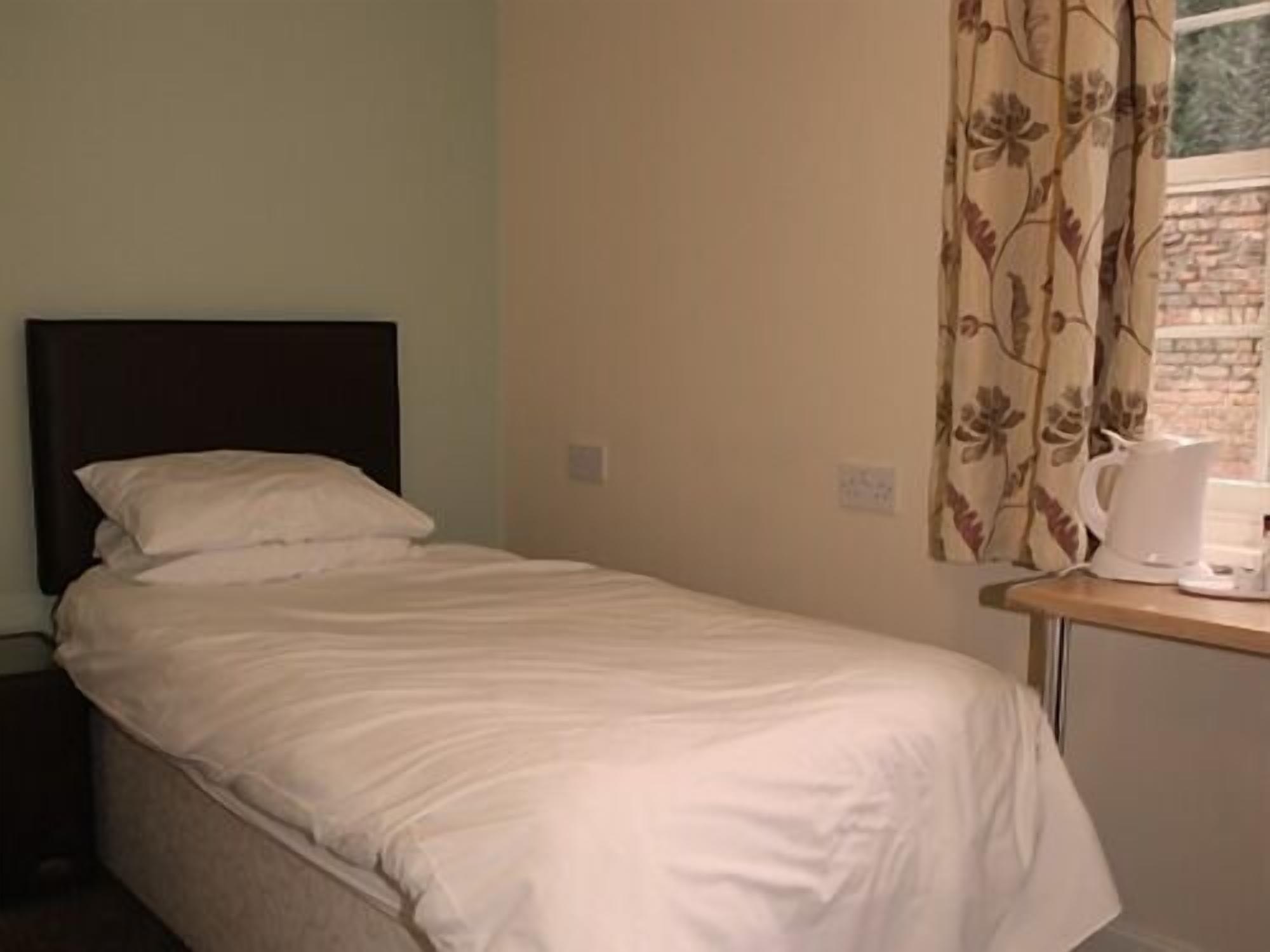 A single bed with a dark headboard, a wooden nightstand with a white kettle, and a window with floral curtains.