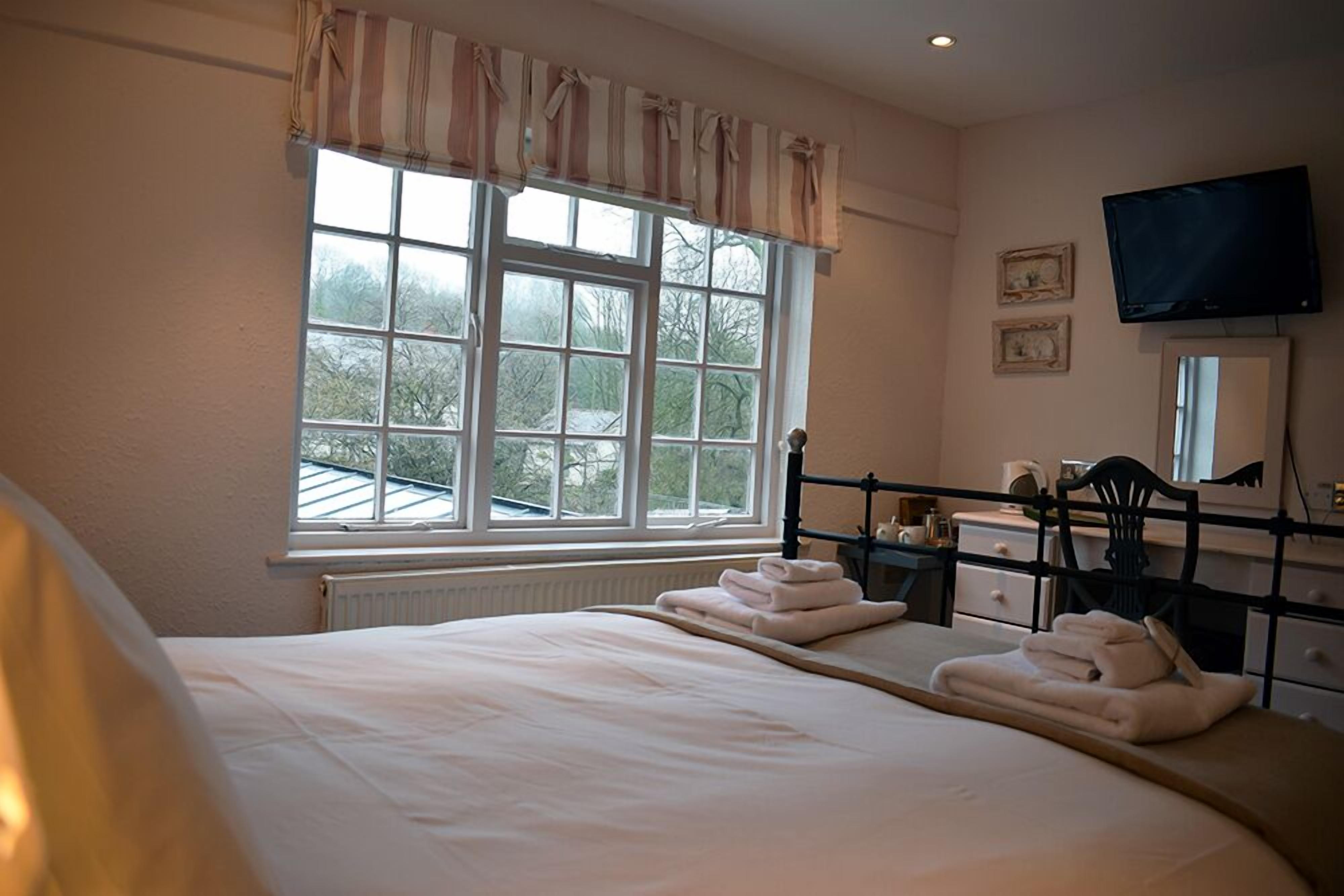 A bedroom with a large bed, a window with curtains, a bedside table, a chair, a dresser, a television, and framed pictures on the wall.