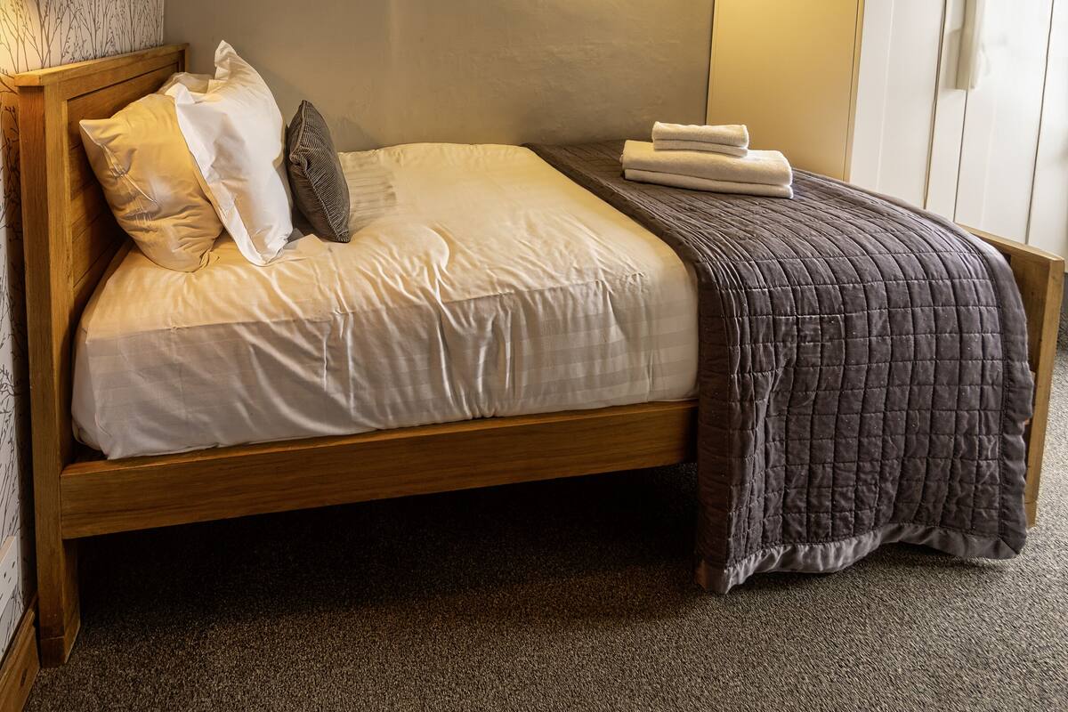 A neatly made bed with a wooden frame, white bedding, and a dark quilted throw.