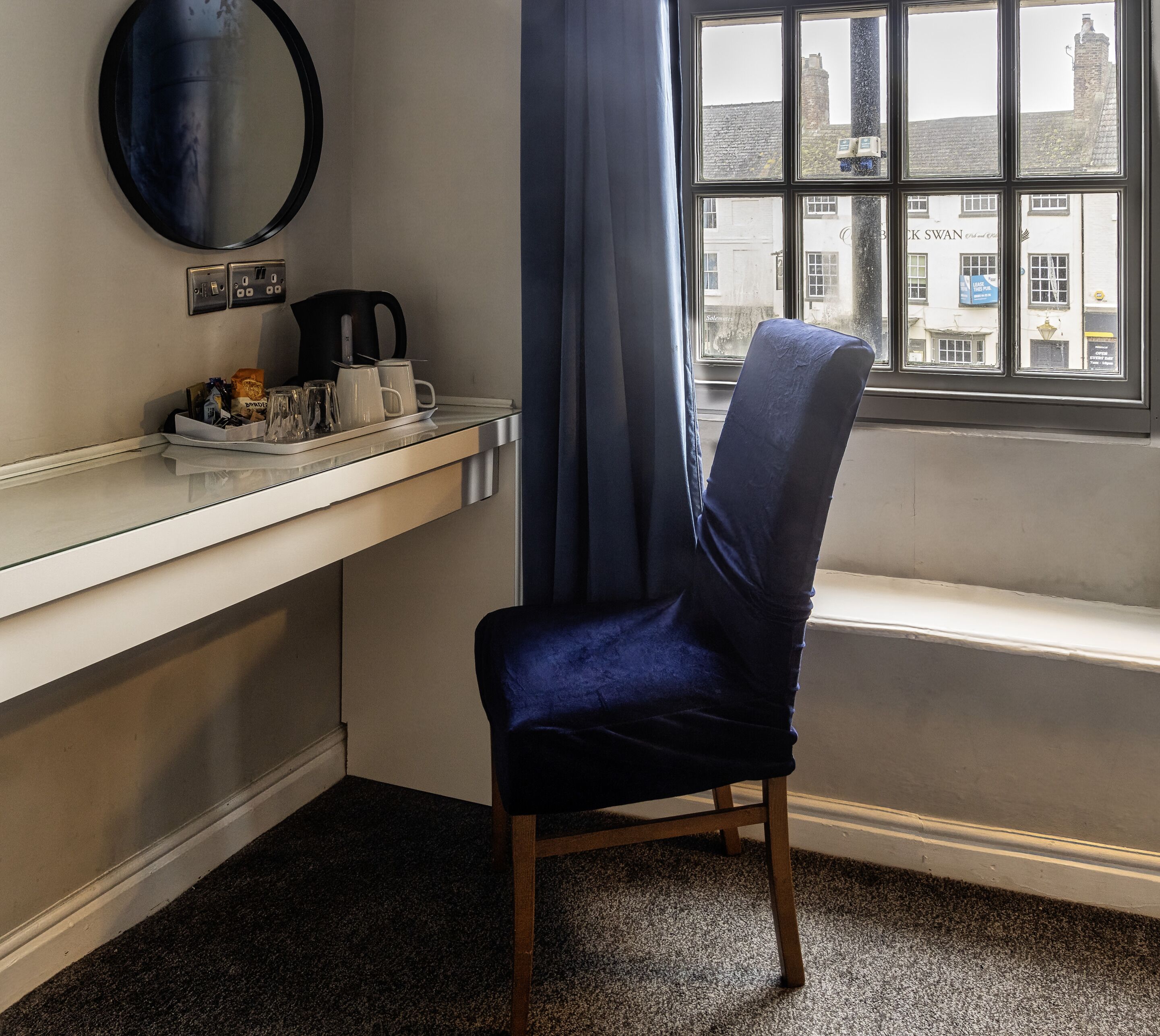 A corner room with a window, a chair, a counter with kitchen items, and a mirror.