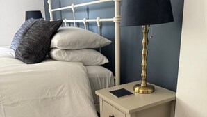 Ein ordentlich bezogenes Bett mit weißen Leinentextilien und zwei Kissen, ein Nachttisch mit Lampe und Buch sowie ein gemustertes Teppich.