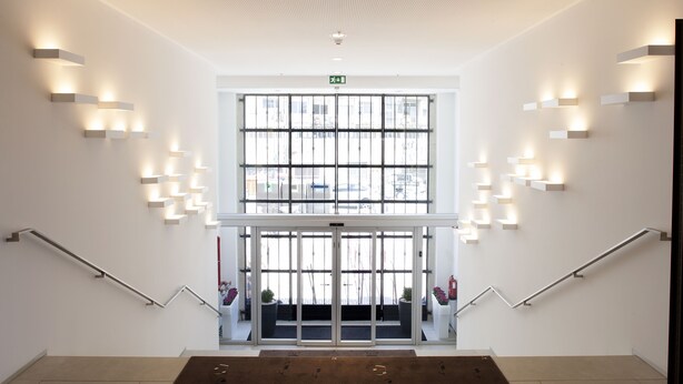 A modern hotel lobby with a central staircase, illuminated wall sconces, and large windows offering views of the city. - Hotel da Música (Porto)