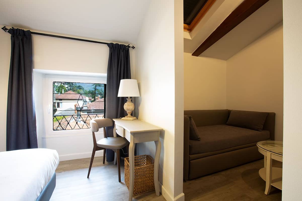 A hotel room with a bed, a desk with a lamp, a chair, a sofa, and a view of the outside.