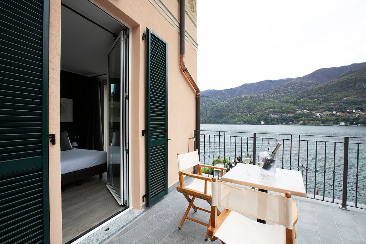 A balcony with a table and chairs, a bed with open shutters, and a view of a lake and mountains.
