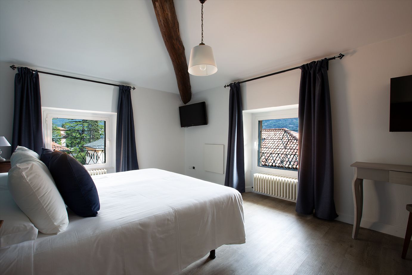 A bedroom with a bed, a TV, a window with curtains, a radiator, and a wooden ceiling beam.