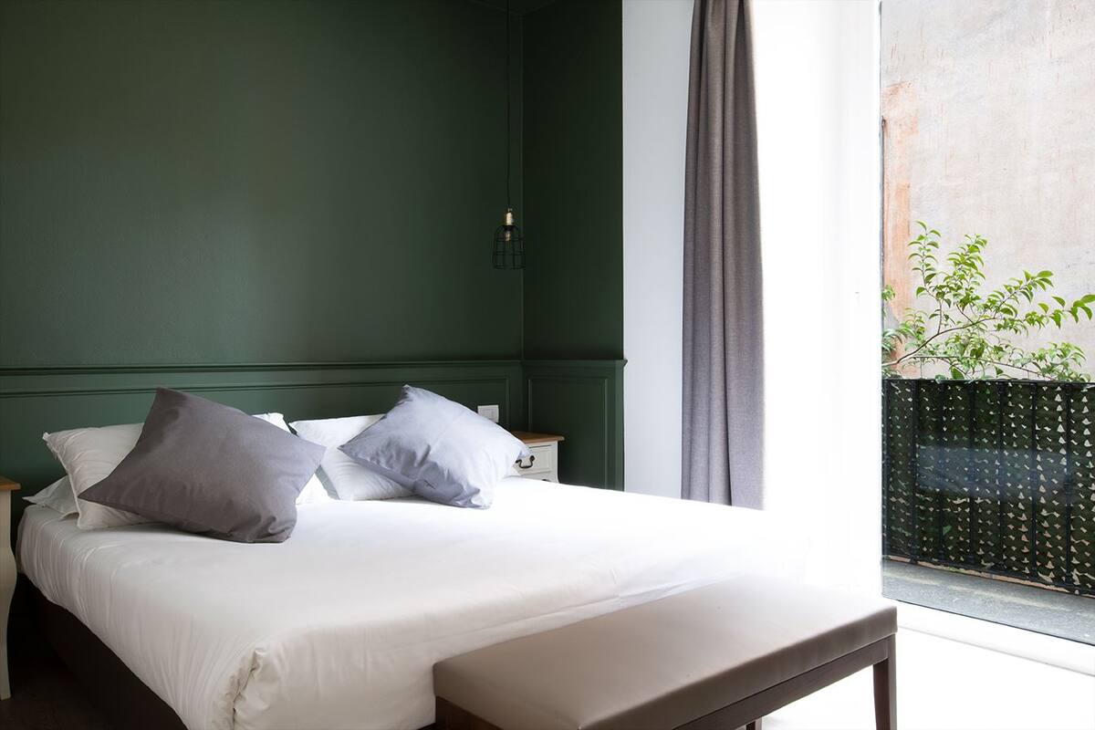 A bedroom with a bed, pillows, a bedside table, a lamp, and a view of greenery outside.