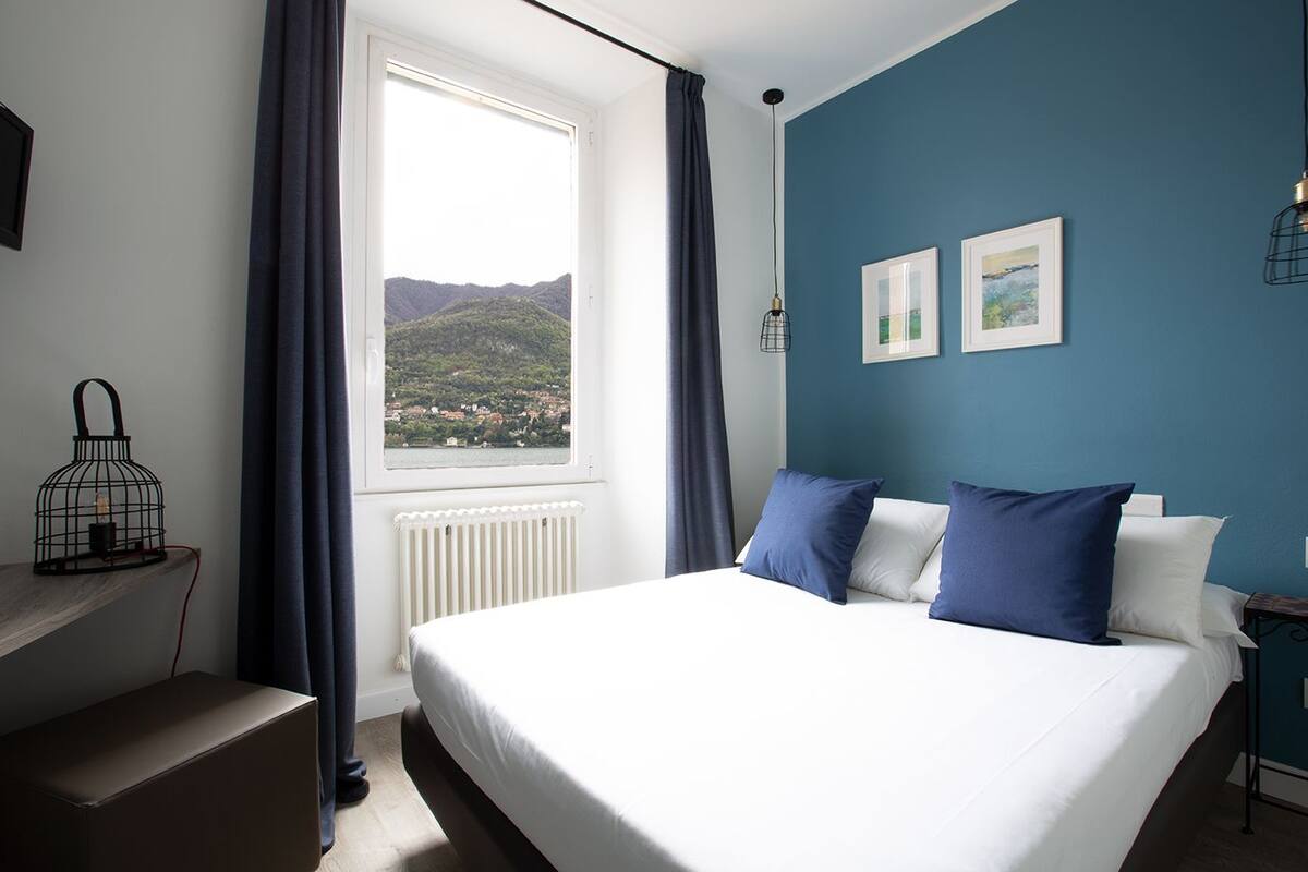 A bedroom with a bed, a window with a view of a mountain, a radiator, and a nightstand with a lamp.