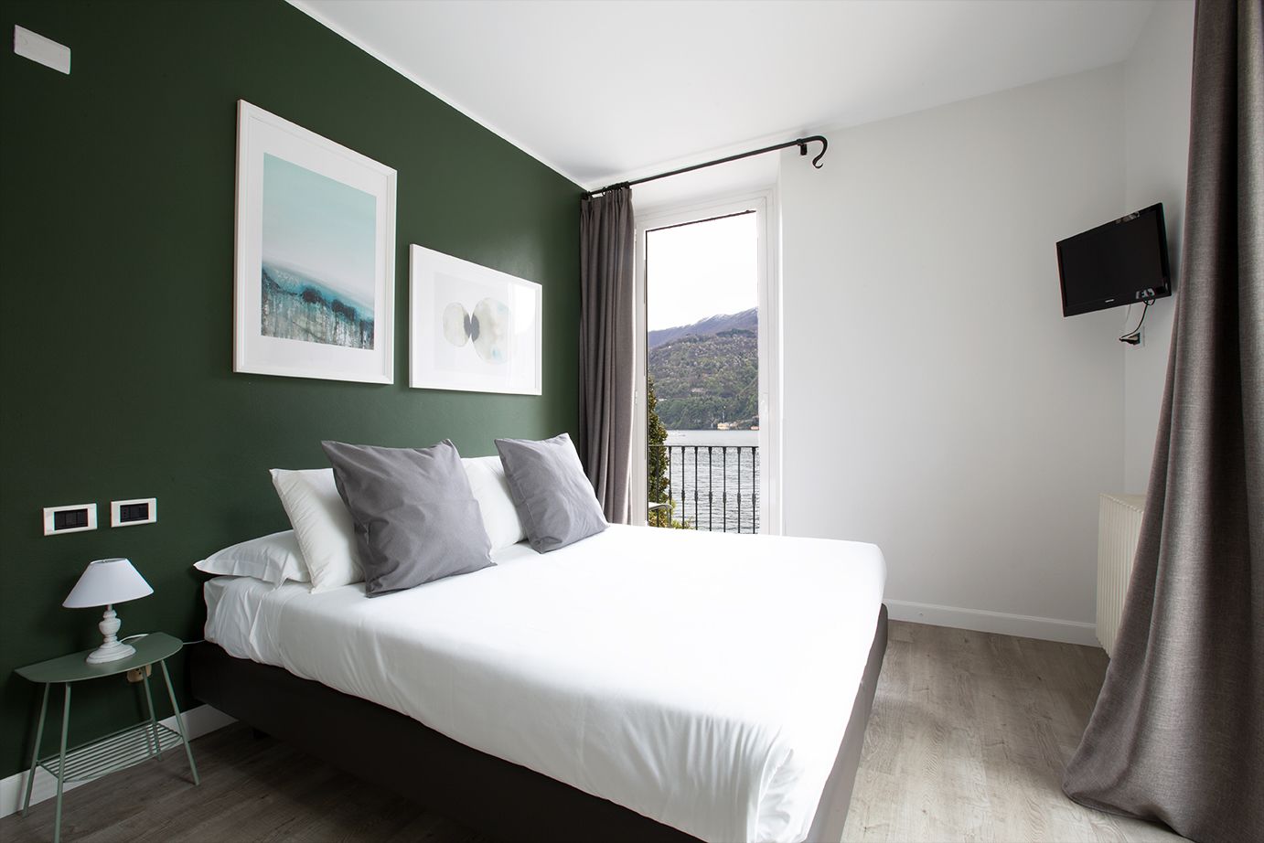 A bedroom with a bed, a nightstand, a lamp, a TV, and a window with a view of a lake and mountains.