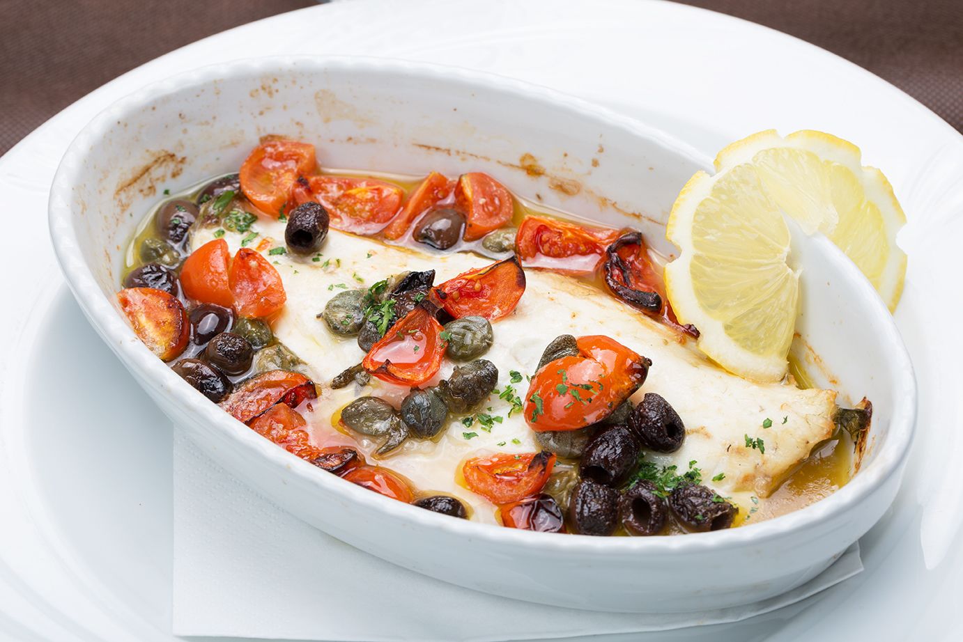 Un plato ovalado blanco con un guiso de mariscos, tomates, aceitunas y rodajas de limón.