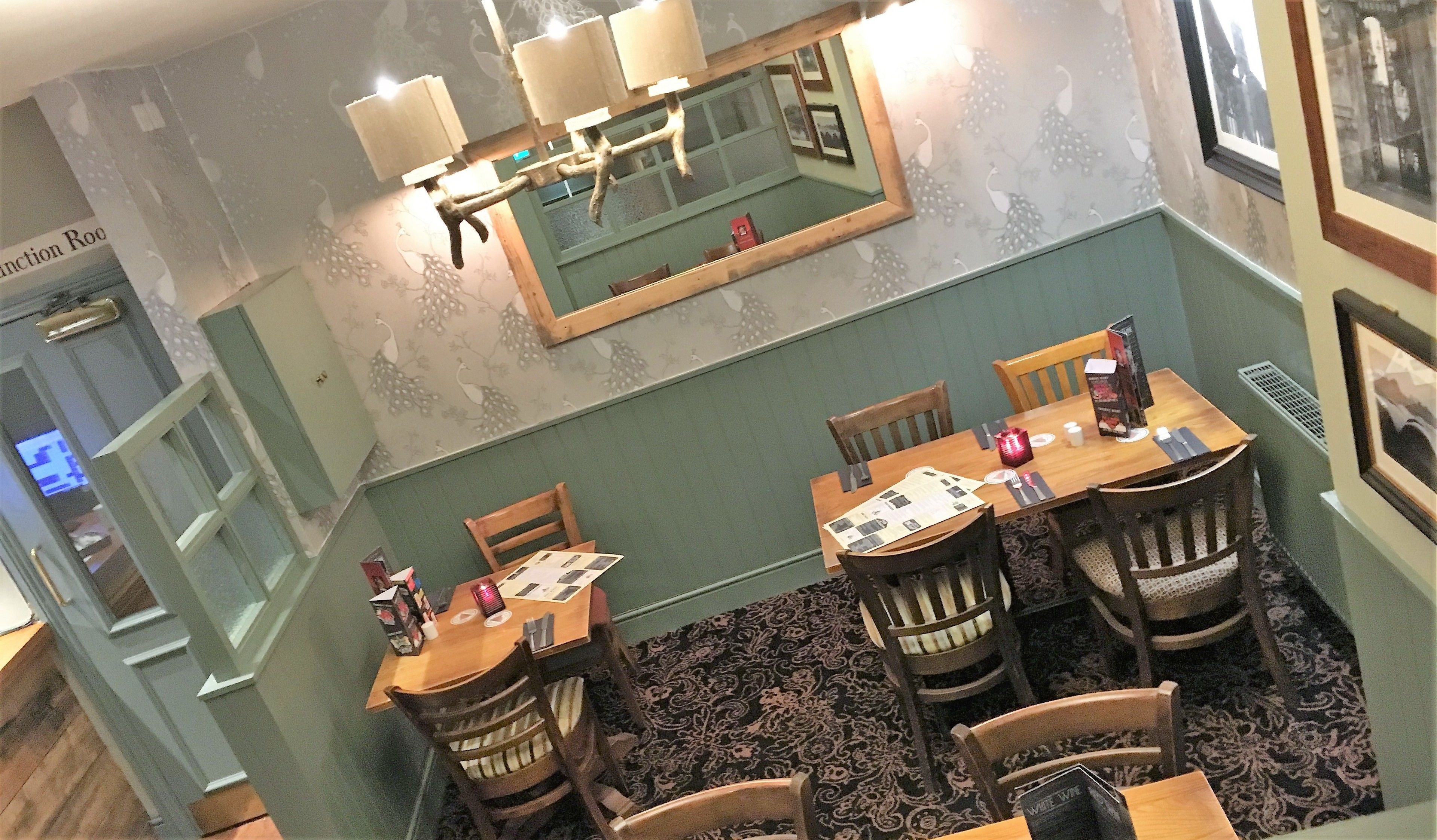 A dining area with a high ceiling, a chandelier, and a large mirror. There are wooden tables and chairs, and a carpeted floor.