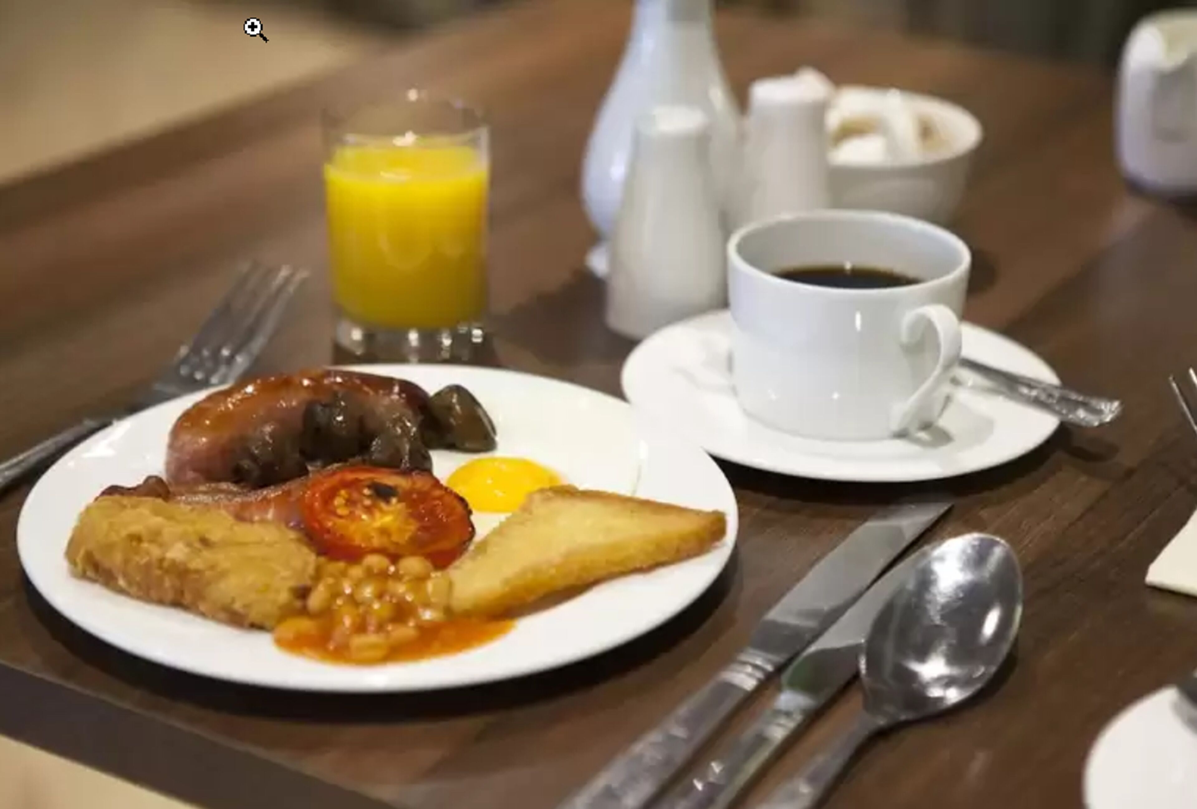 Un plato de desayuno con huevos, frijoles, tomates y tostadas, un vaso de jugo de naranja y una taza de café sobre una mesa de madera.
