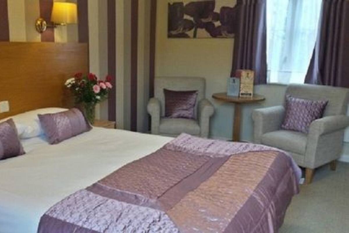A hotel room with a large bed, two armchairs, a small table, and a nightstand with a lamp.