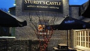 A stone building with a sign reading "Sturdy's Castle" indicating it is a pub, restaurant, and hotel. There are outdoor tables and umbrellas, and a tree in the center. - Sturdy's Castle Country Inn (Kidlington)