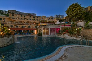 A swimming pool with a stone border, a covered area with a glass wall, and a building with balconies in the background.