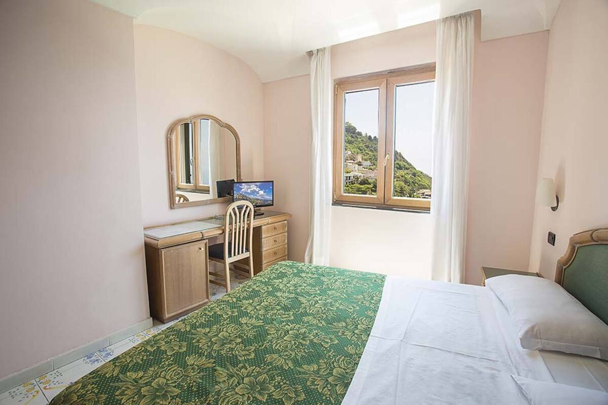 A hotel room with a bed, a desk with a chair, a television, and a window with curtains.
