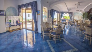 Una hall d'albergo con pavimento piastrellato, tavoli rotondi, sedie in vimini e vista sull'esterno.