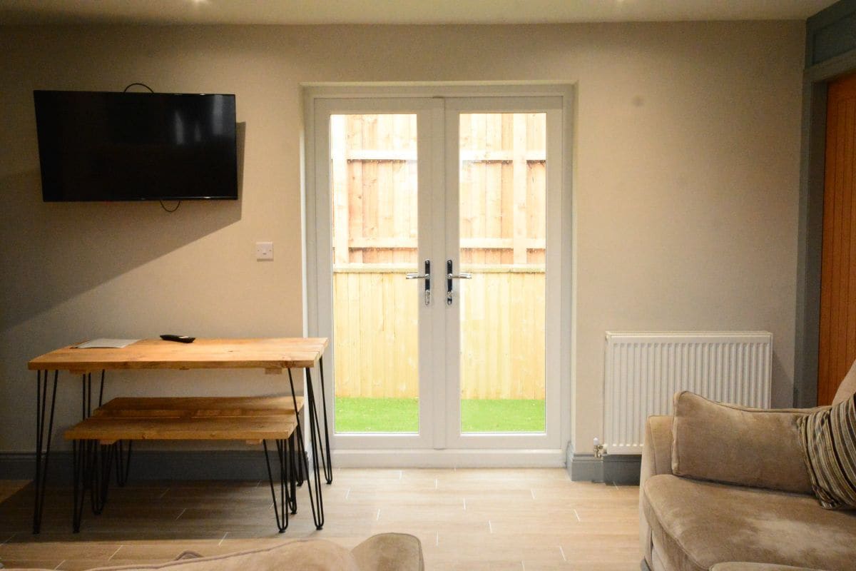 A living room with a TV mounted on the wall, a wooden table with benches, a sofa, and a sliding glass door leading to a backyard.