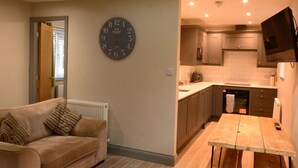 A compact living space with a sofa, a dining table, a kitchen area with cabinets and a TV, and a clock on the wall. - Durham City Cottages (Durham)