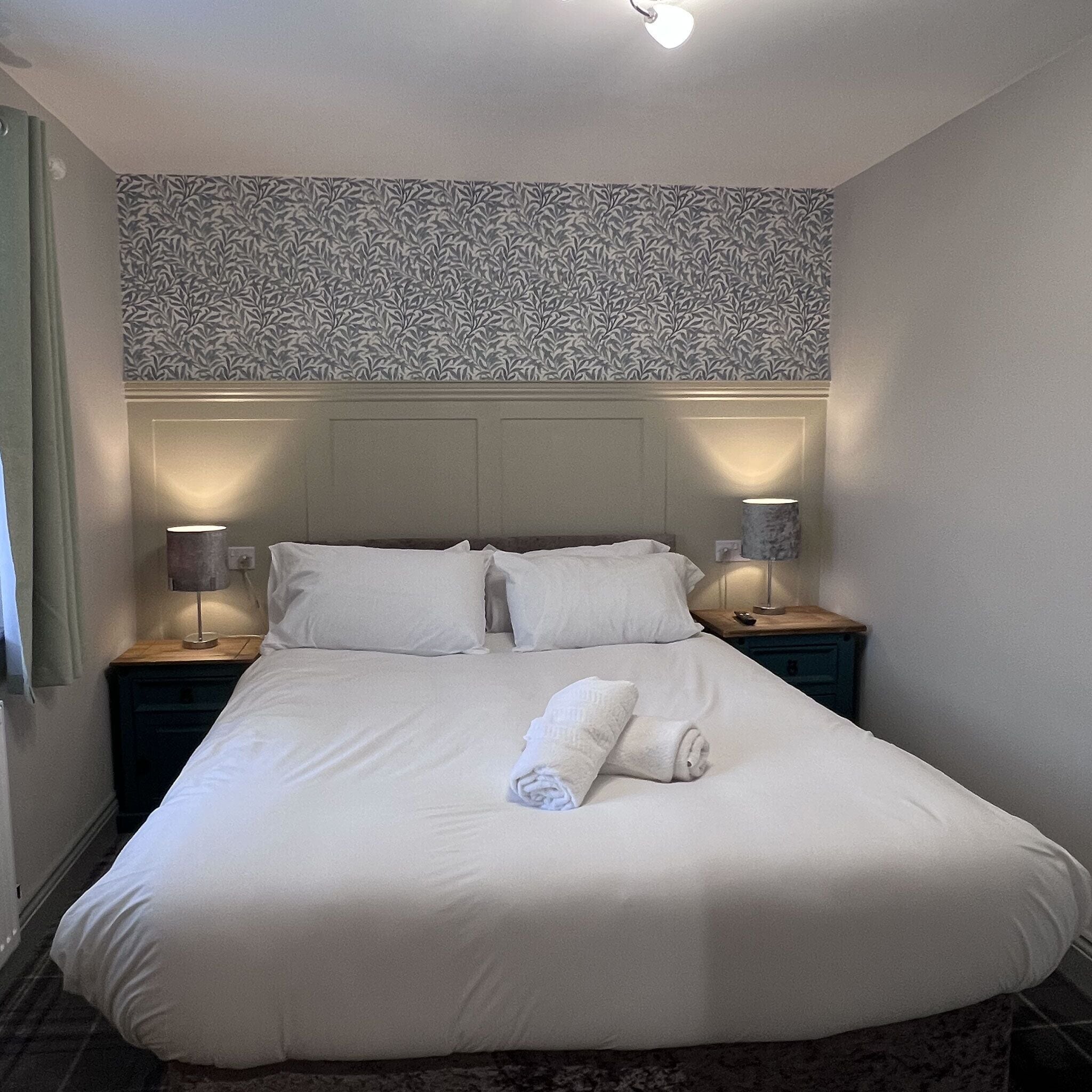 Un lit bien fait, avec du linge de lit blanc et une serviette roulée, deux lampes de chevet, un papier peint à motifs et un mur uni.