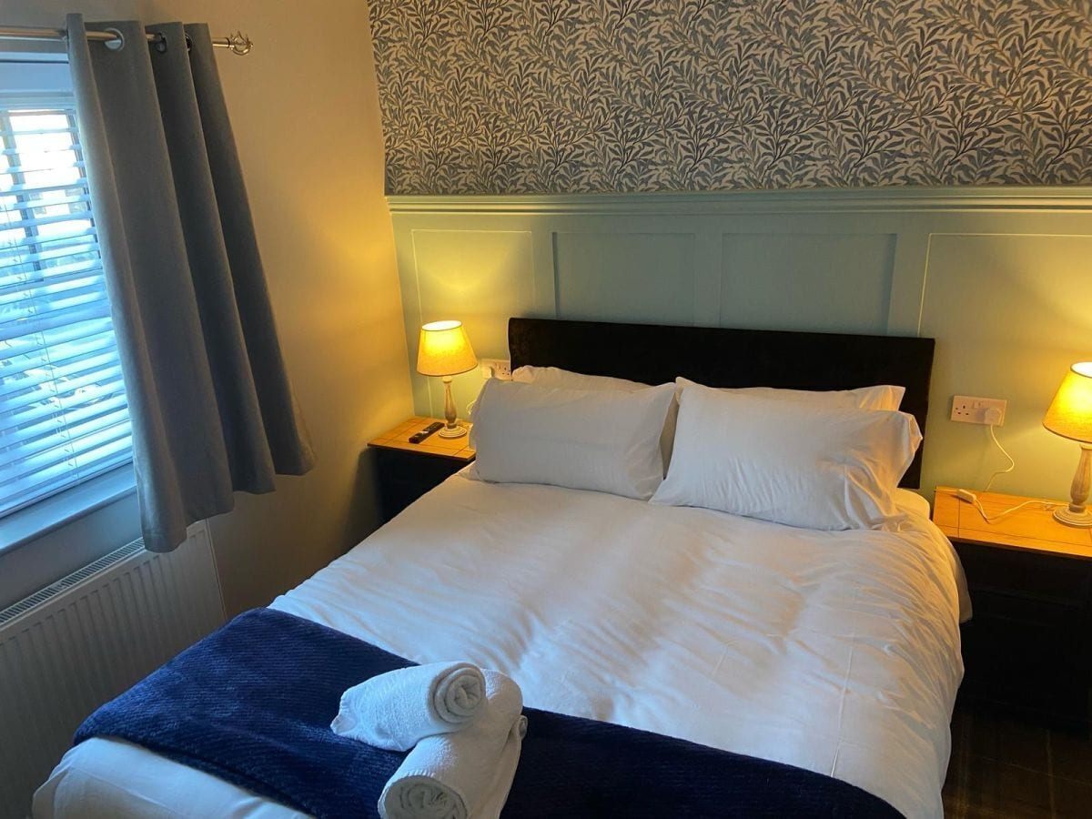 Un lit bien fait, avec du linge de lit blanc et un plaid bleu, deux lampes de chevet et une fenêtre avec des rideaux.