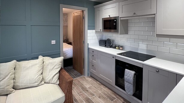 A compact kitchen with a microwave, stove, and white tiled backsplash. A wicker sofa is positioned near a doorway leading to a bedroom. - Durham City Cottages (Durham)
